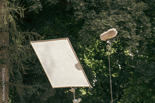 Obraz na płótnie A large rectangular diffusion light reflector panel and a fur-covered boom microphone stand outdoors against a dense background of green trees during a film set production.