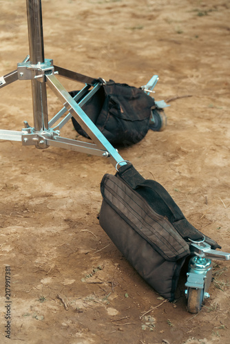 фотографія A professional metal light stand base equipped with caster wheels and weighted black sandbags sits on dusty ground at an outdoor film location