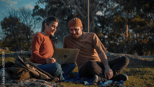 Young Latino couple sitting in a park looking at a laptop together, highlighting digital connection, companionship, collaboration, and a modern outdoor lifestyle.