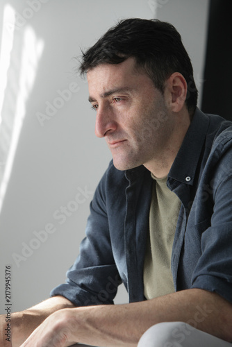 Canvastavla Thoughtful and pensive mid adult man sitting in natural window light, showing a quiet and reflective mood with signs of tiredness, sadness, stress and mental burnout