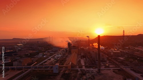 Massive industrial factory complex bathed in the warm golden light of setting sun. Multiple tall smokestacks rise from the facility, releasing plumes of white smoke and steam.