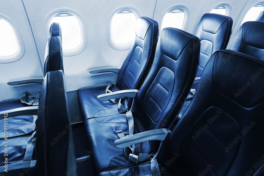 Obraz premium Empty blue passenger seats line a commercial airplane cabin beside bright windows. Seat belts armrests and leather upholstery fill the aircraft interior for passenger travel background.