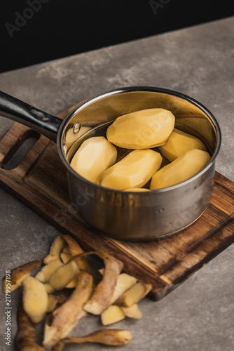 Peeled raw potatoes in a stainless steel saucepan placed on a wooden cutting board with potato skins lying nearby on a stone kitchen countertop