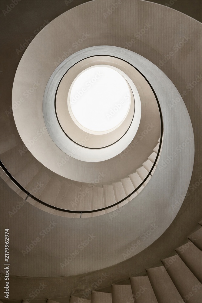 Fototapeta premium Spiral staircase viewed from below with round skylight at top. Minimalist concrete architecture with geometric circular shapes and natural light.