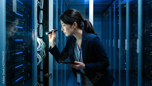 Naklejka 青い光のデータセンターでラックの配線を懐中電灯で照らし点検する女性技術者の横顔、タブレットパソコンと配線ケーブルも見える