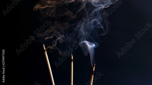 Close-up of three burning incense sticks releasing delicate curling smoke against a deep black background, thin aromatic trails forming abstract shapes in darkness, Antyesti, Antim