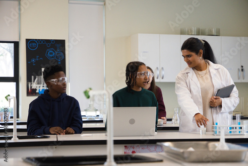 Diverse teens watching Indian instructor in lab coat pointing at test tubes with tablet copy space