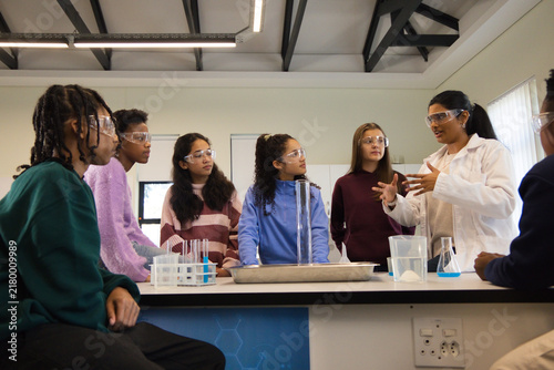 Diverse teenage students in lab watching Indian instructor in lab coat showing with tubes, goggles
