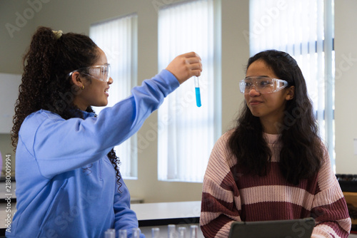 Two African American teen girls wearing goggles examining blue test tube using tablet at school lab
