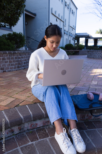 Fotografía Asian girl sitting on brick ledge in courtyard using laptop, wearing sweater and