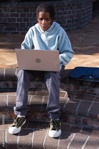 Bild auf Leinwand African teen male sitting on step in brick courtyard wearing hoodie with blue ba