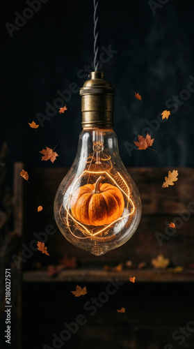 Pumpkin inside glowing vintage light bulb surrounded by floating autumn leaves, creating warm and cozy fall atmosphere with rustic background