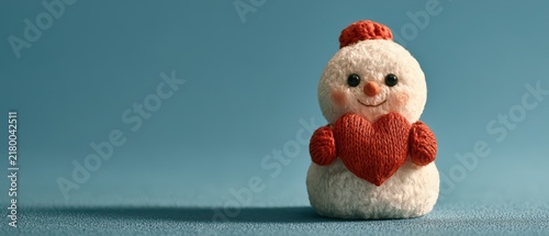 Cute small handmade snowman holding red heart knitted decoration on blue textured background for winter love
