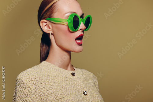 Canvastavla Stylish woman in green sunglasses and beige textured jacket looks surprised with open mouth on plain background