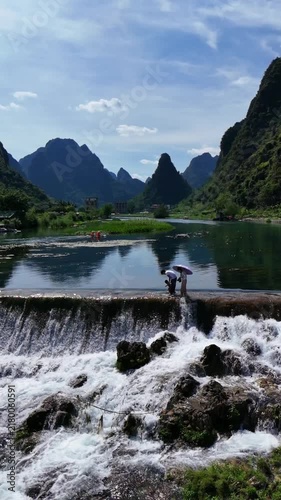 Vertical Video: Slow-motion view of a family walking across a stone dam with calm water above and a white waterfall cascading below