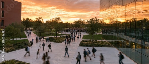 Students walking on university campus plaza at sunset with glass building and orange sky