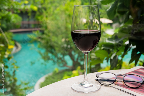 Glass of red wine, book, and glasses on a table overlooking a pool and garden.