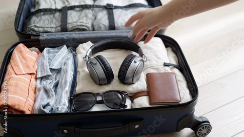 Woman hand packing open suitcase with clothes, wireless headphones, sunglasses, and wallet on light floor, travel preparation, organized luggage, vacation concept, close up shot