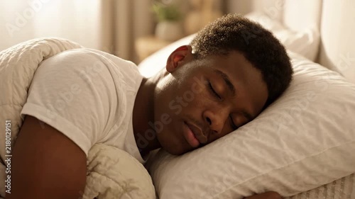 Young African American man sleeping peacefully in cozy bed on soft pillow under blanket, morning sunlight, healthy rest, deep sleep quality, relaxation, bedroom interior, well-being concept shot