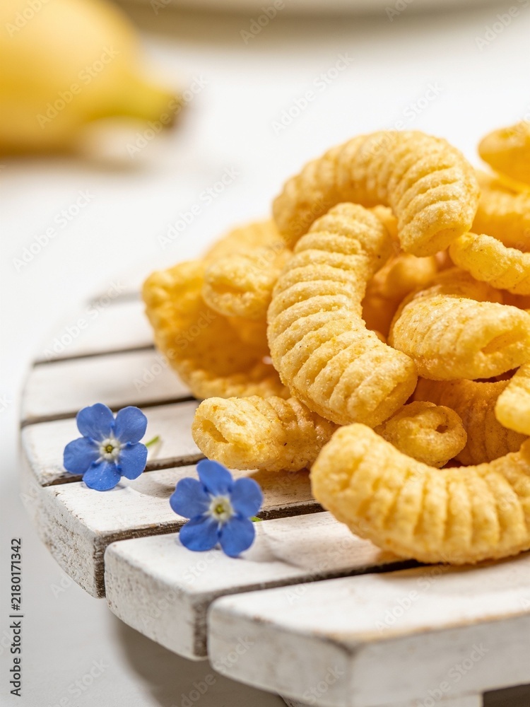 Obraz premium A delightful close-up shot of crispy golden corn puffs, beautifully piled on a rustic white wooden pallet coaster. Adorned with tiny, delicate blue flowers that add a poetic natural contrast.