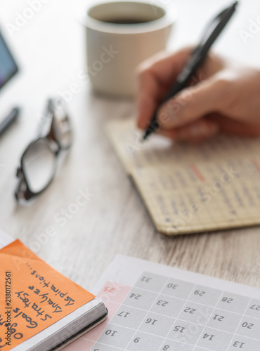 Person's hand writing notes in a notebook, managing a schedule and planning goals, tasks, and responsibilities for productivity on a wooden desk with a calendar and coffee
