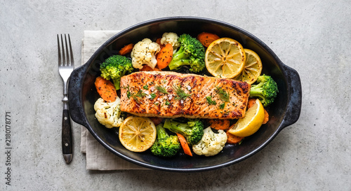 Top-down view of a seasoned grilled salmon fillet served with roasted broccoli, cauliflower, carrots, and lemon slices in a black dish with a fork.