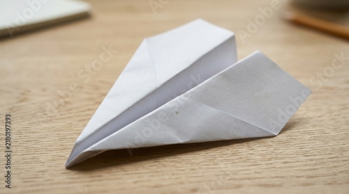 Origami paper airplane lies diagonally on a textured light wooden desktop with blurred background elements.