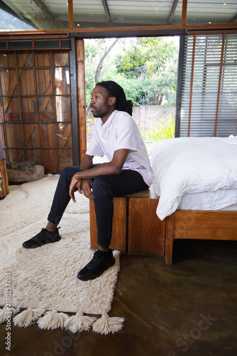 Foto African man sitting on wooden bench in bedroom, gazing through large glass windo