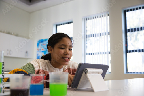 Child girl student wearing knit sweater sitting in classroom using tablet with colored cup liquids