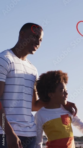 Animation of hearts over african american father and son standing on beach at sunset