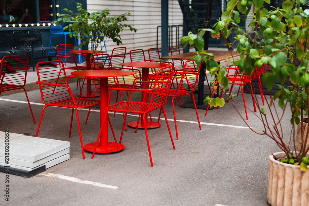Obraz premium A row of red chairs surrounds several circular tables in an outdoor seating area. The space features potted plants and is set on a paved surface in a city environment