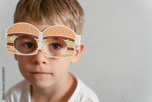 A young boy wearing creative novelty paper glasses shaped like hamburgers, looking directly at the camera with a neutral yet playful expression in an indoor setting.