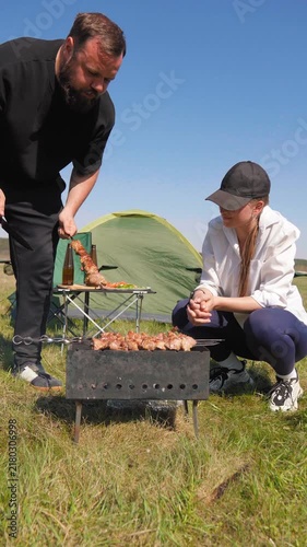 Young man and woman enjoying a picnic in nature on a sunny day, with the man turning skewers of shashlik on a charcoal grill while the woman watches next to their camping tent