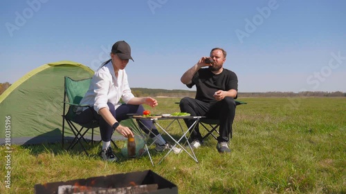 Young man and woman sitting on camping chairs, drinking beer, and enjoying a picnic near a tent and a barbecue grill on a sunny day in a beautiful green field