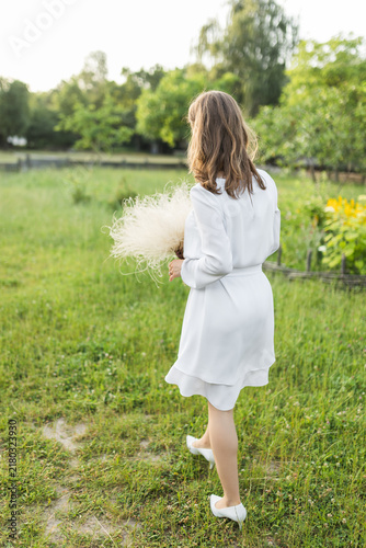 Фотография Bride in short white dress holding dry bouquet walking on green grass meadow