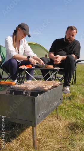 Young man and woman grilling meat and having a picnic in a green field during a sunny day, sitting in camping chairs next to their tent and enjoying food and drinks in the great outdoors