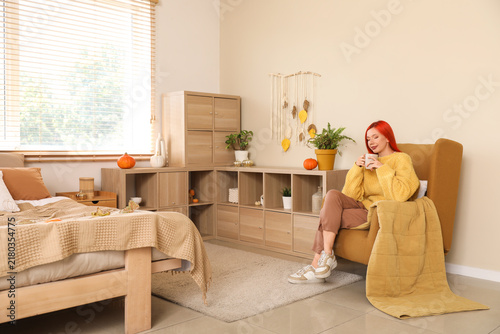 Beautiful young happy woman with autumn decor and cup of tea sitting on armch...