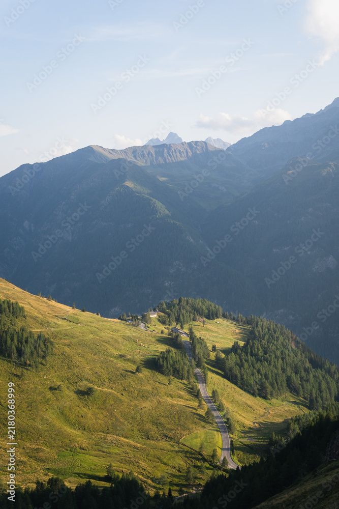 Obraz premium Alpine Mountain Landscape with Winding Asphalt Road Illuminated by Evening Sunlight