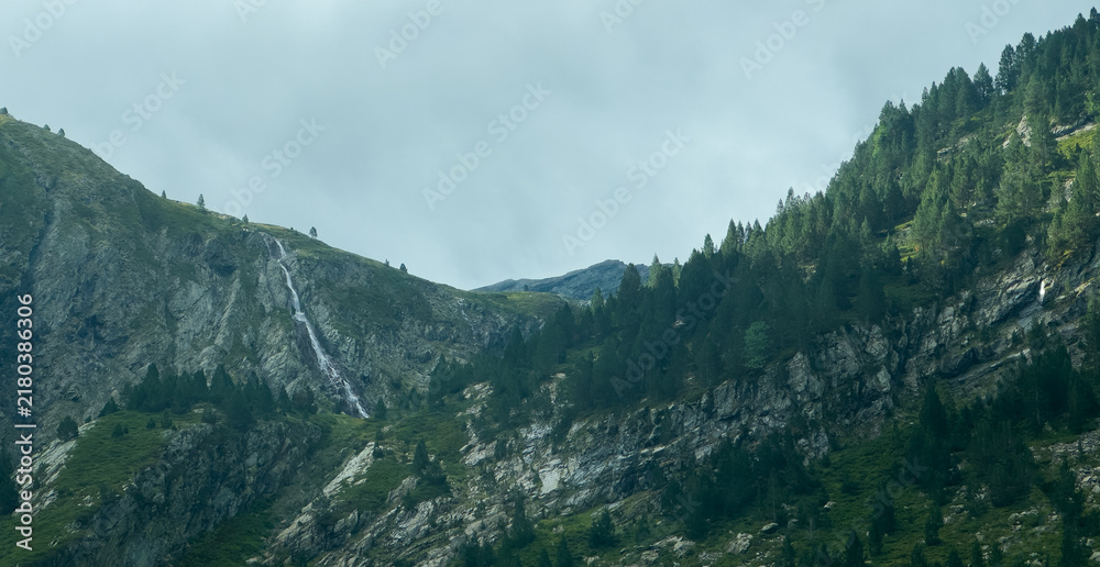 Obraz premium serene mountain landscape with cascading waterfall and rugged terrain under a cloudy sky, aerial perspective showing evergreen trees and rocky formations in a natural setting.