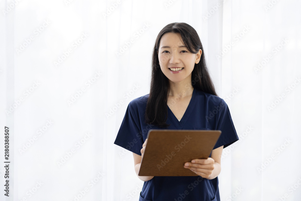 Obraz premium Young Japanese female doctor in scrubs holding a clipboard and smiling at the camera by a bright window