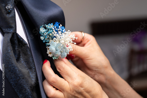 Close up of female hands pinning a blue and white dried flower boutonniere onto the navy lapel of an elegant groom suit, tender detail of wedding day preparation with red nails and a gold ring