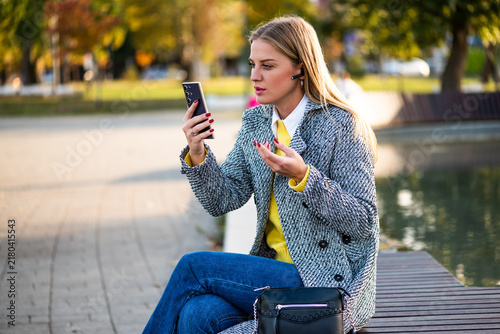 Confident and urban businesswoman sitting outdoor in autumn, using smartphone and headphones for professional video call in the city park.