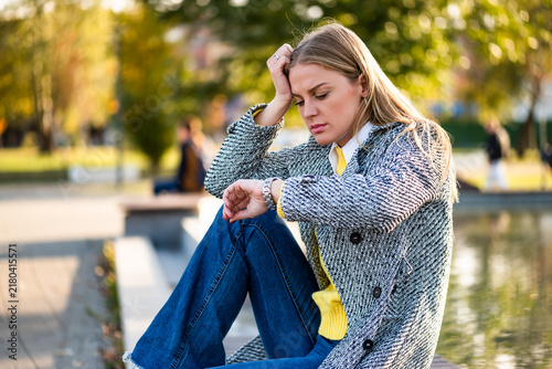 Stressed professional woman sitting in a public park, looking at her watch with frustration and anxiety while waiting and worrying about time.