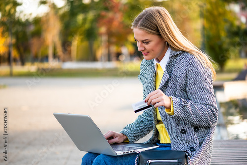 Freelance businesswoman using laptop in the city park while managing online purchases and financial activities on a sunny autumn day.