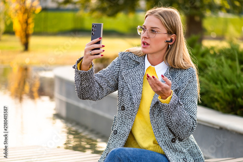 Stylish businesswoman with eyeglasses working remotely outdoor, using smartphone and headphones to stay connected during an online meeting on a autumn day.