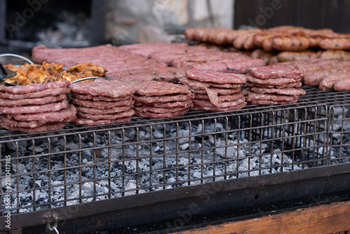 Stacks of raw burger patties, sausages and chicken piled on a metal barbecue grill over grey embers and charcoal, rustic outdoor cooking preparation for a summer party celebration