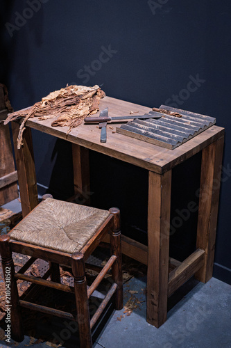 Vintage cigar rolling workstation featuring dried tobacco leaves, cutting tools, a wooden molding board, and a rustic straw-seat stool.
