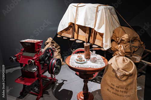 Historical cocoa processing exhibition featuring a restored red mechanical grinder, jute sacks, wooden crates on a cart, and a small table with a copper chocolaterie.