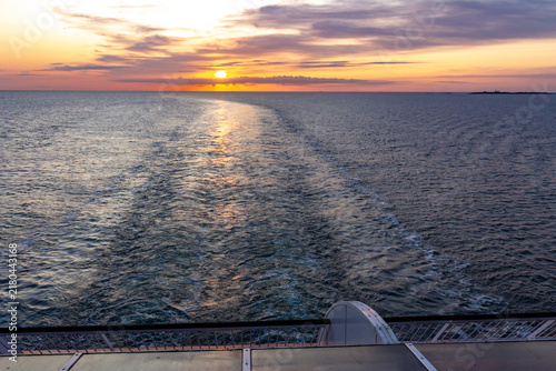 Картината върху платно A wake trail left by a cruise ship or ferry on the calm sea surface during a golden sunrise or sunset, viewed from the upper deck