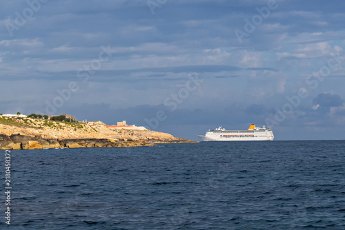 White cruise ship sailing beside rocky coastline across dark blue sea, dramatic layered clouds creating scenic maritime travel atmosphere